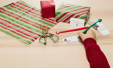 Load image into Gallery viewer, Person wrapping a gift with red and green striped paper, scissors, and a pen on a wooden surface.