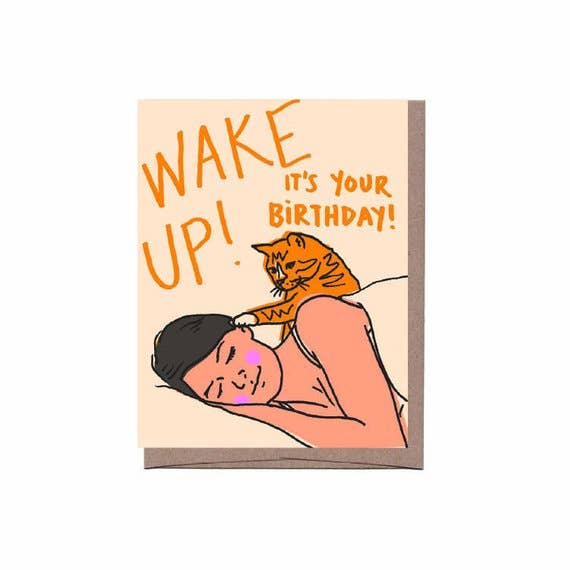a crem coloured crd with an illustration of a woman asleep i her bed with an  tabby  cat touching her face with its paw as ifto be waking her up paired with a brown kraft envelope