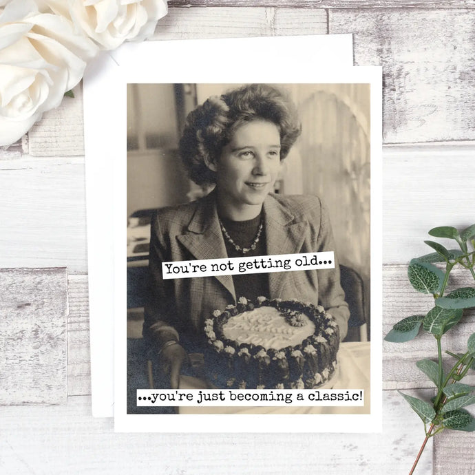 a black and white photo on a greeting card of a woman in front of a vintage 50's birthday cake. text. you're not getting old...you're just becoming a classic!