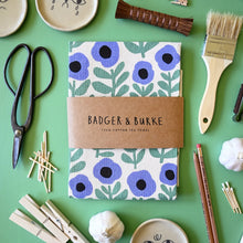 Load image into Gallery viewer, A flat-lay of a Badger &amp; Burke cotton tea towel with a blue and green floral pattern, wrapped in a brown kraft paper band. The towel is styled on a mint green background with various craft and kitchen props including scissors, paintbrush, garlic, and wooden clothespins.
