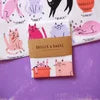 Load image into Gallery viewer, A small pink and white patterned card or booklet with a gold border, placed on a purple surface. The card has a pink illustration of a woman's face and hair at the top and a small pink graphic at the bottom.
