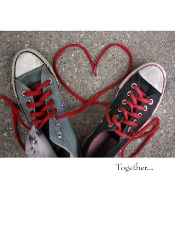 a colour photo of a grey converse sneaker and a black converse sneaker chuck taylors with red she laces in each intertwined making a big red hearth on the pavement 