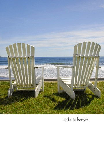 a colour photo of 2 anarondack chairs side by side facing the ocean or a big lake shore on green grass with blue water . text says life is better