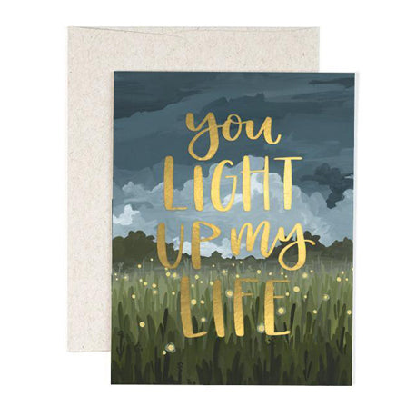 a colour illusrtation of a grass feild and clouds in sky with light dots appearing to be fireflies in green and dark blues with gold script you light up my life paired with a light grey envelope 