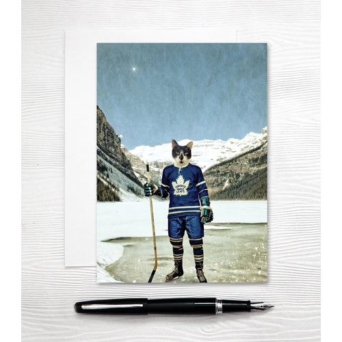 a colour phot of a blak and white cat wearing a toronto maple leafs hockey uniform in skates with a stick on a frozen lake with large mountains in background 