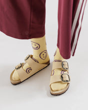 Load image into Gallery viewer, a pair of feet wearing sandles and yellow butter coloured baggu socks with purple coloured smiley or happy faces on them

