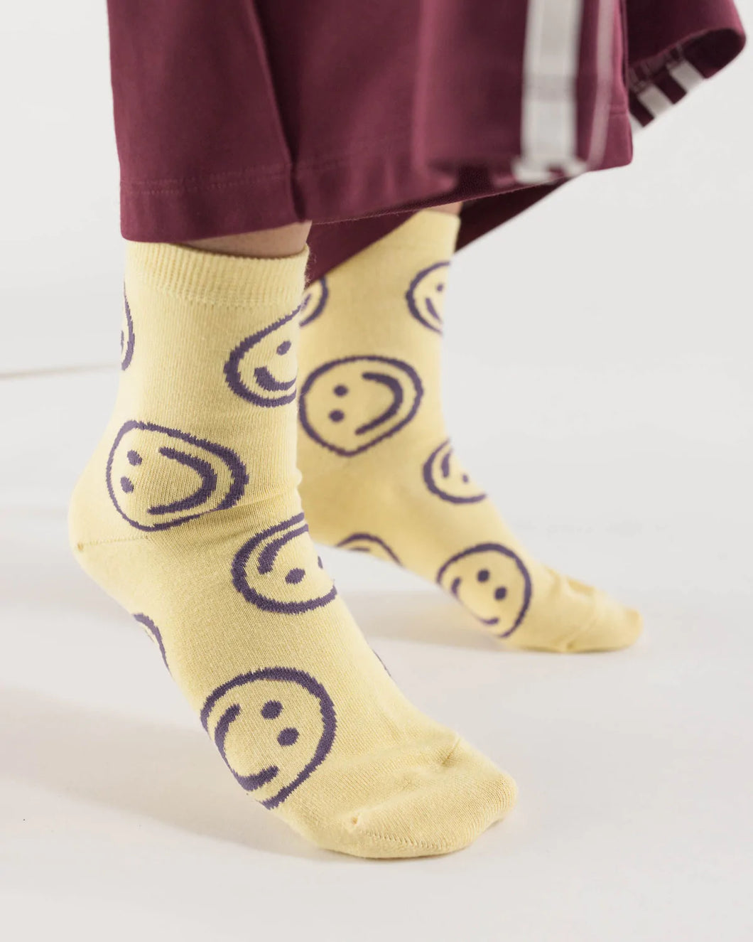 a pair of feet wearing butter yellow coloured socks with purple happy or smiley faces all over them by baggu