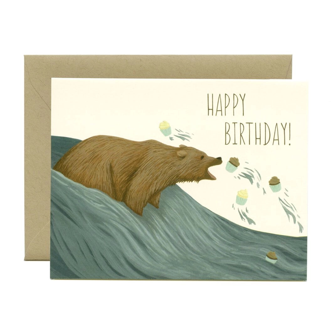 a colour illustration of a grizzly bear in a water fall catching the cupcakes as they swim up stream . text happy birthday 