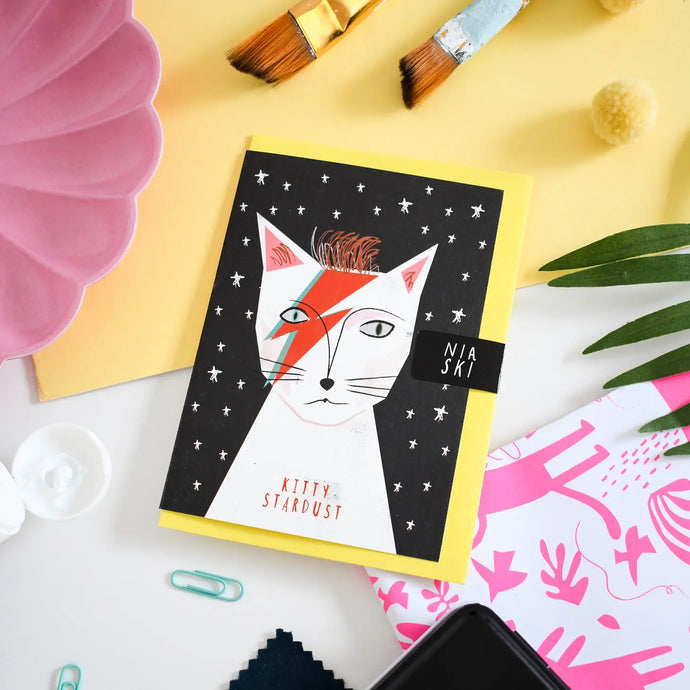 a greeting card featuring a cat that looks like david bowie character ziggy stardust with red makeup on. text. kitty stardust 
