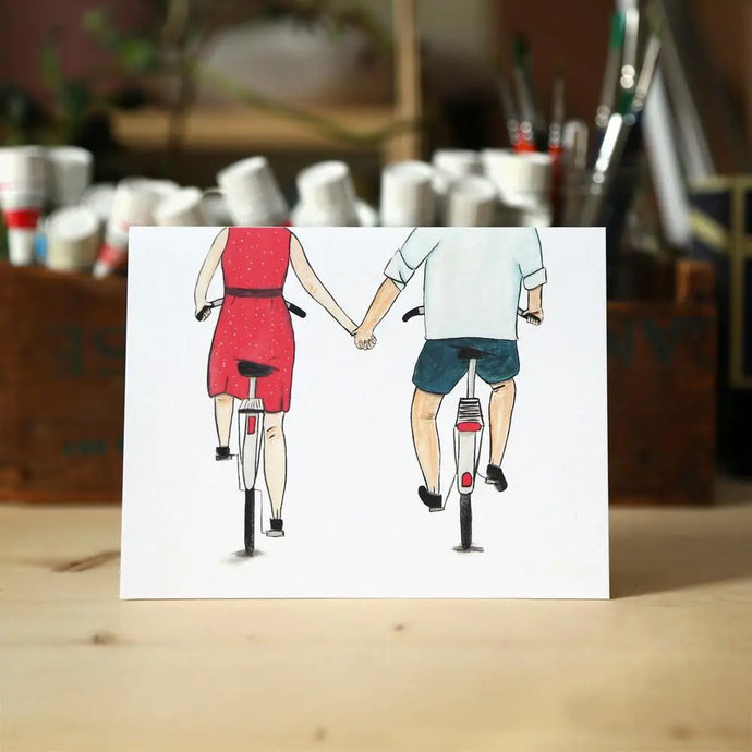 a greeting card with an illustration of a man and woman riding separate bicycles holding hand 